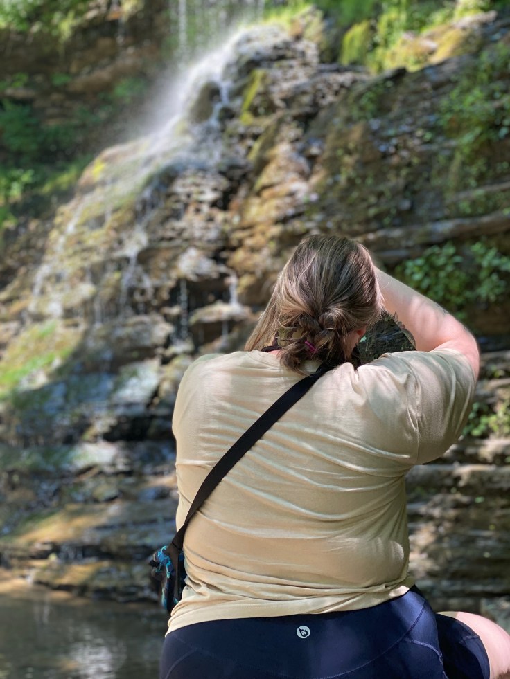 A woman with her back to the camera, aims her own camera at a tall waterfall in the background.